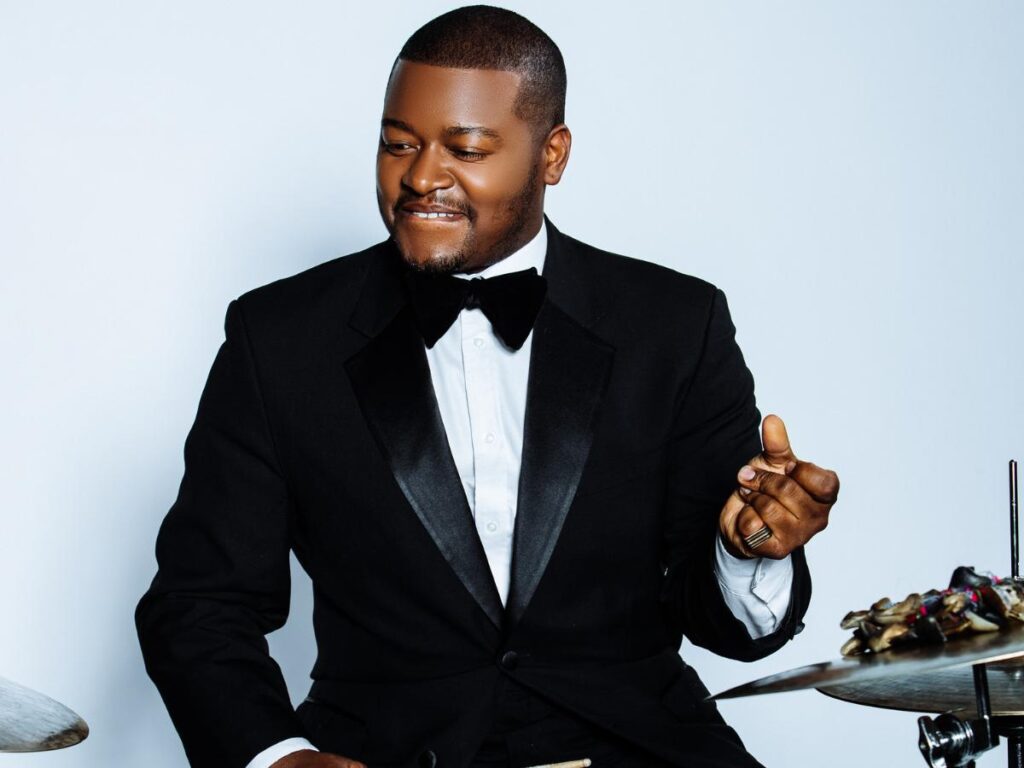 Dante Pope plays the drums while wearing a tuxedo in front of a white backdrop