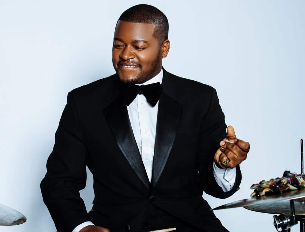Dante Pope plays the drums while wearing a tuxedo in front of a white backdrop