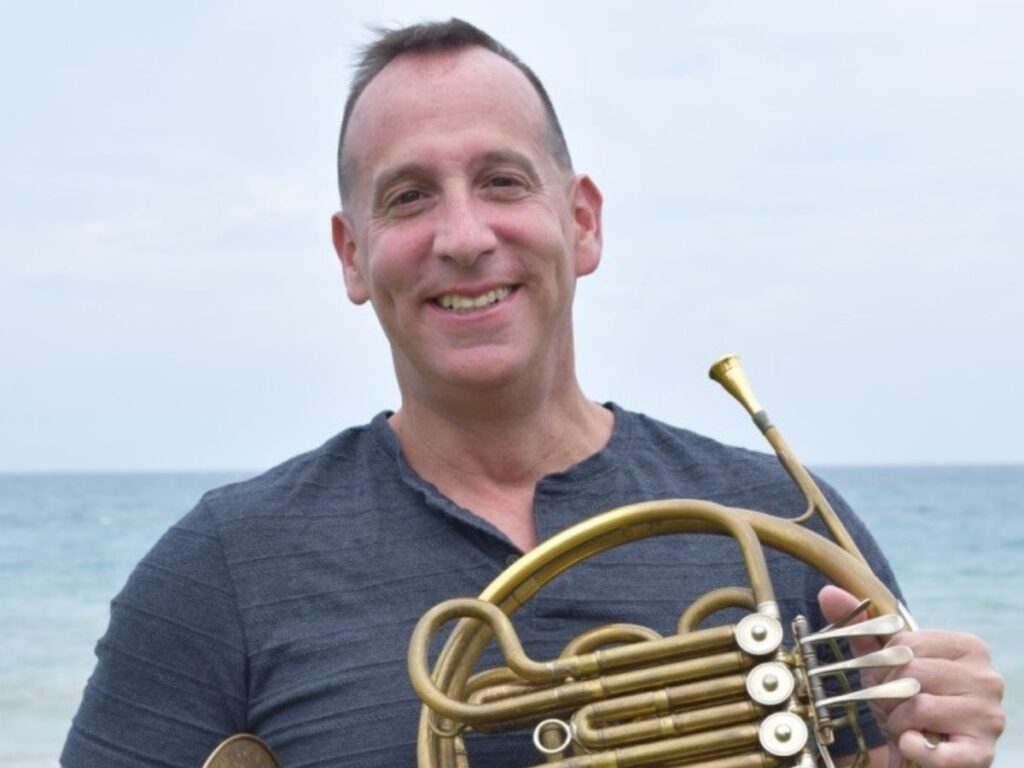 Neal Robert Abarbanell stands on the beach with his french horn, smiling