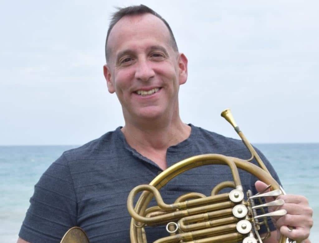 Neal Robert Abarbanell stands on the beach with his french horn, smiling