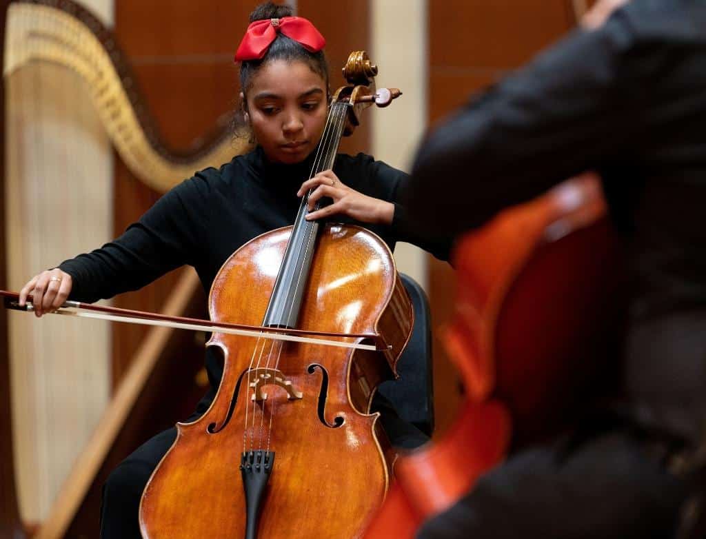 A cello student with a red bow in their hair plays the cello on stage