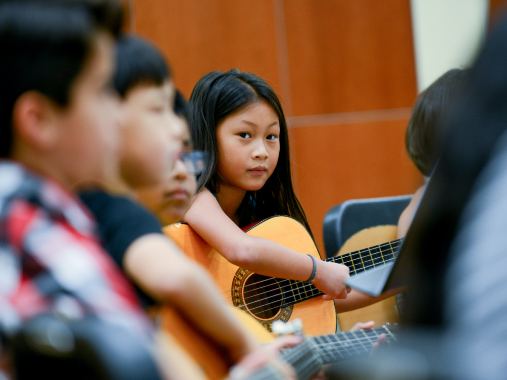 Several children playing guitar, with a young girl in focus holding her guitar and looking at the camera.