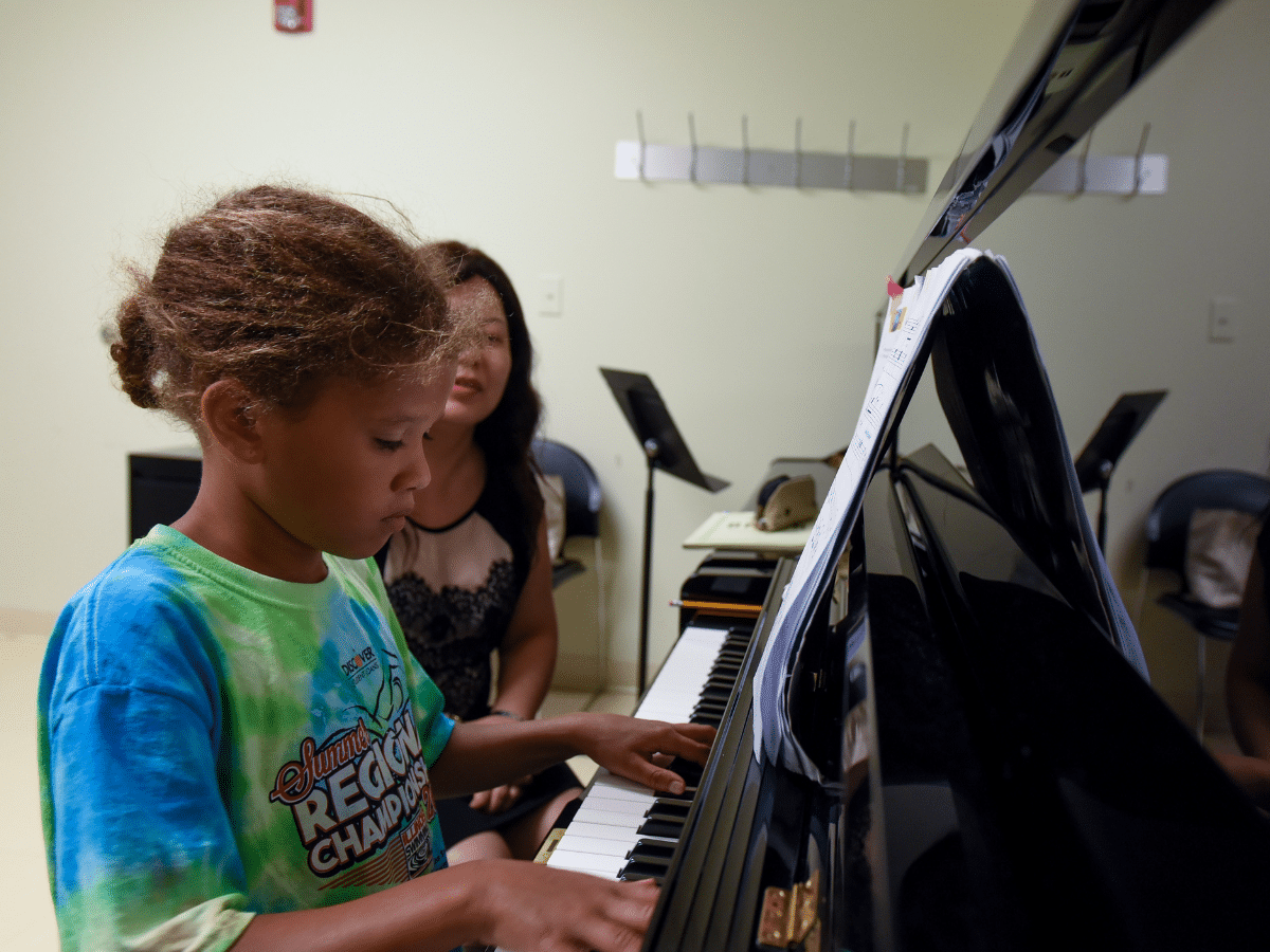 A young student playing piano with her teacher in the background.
