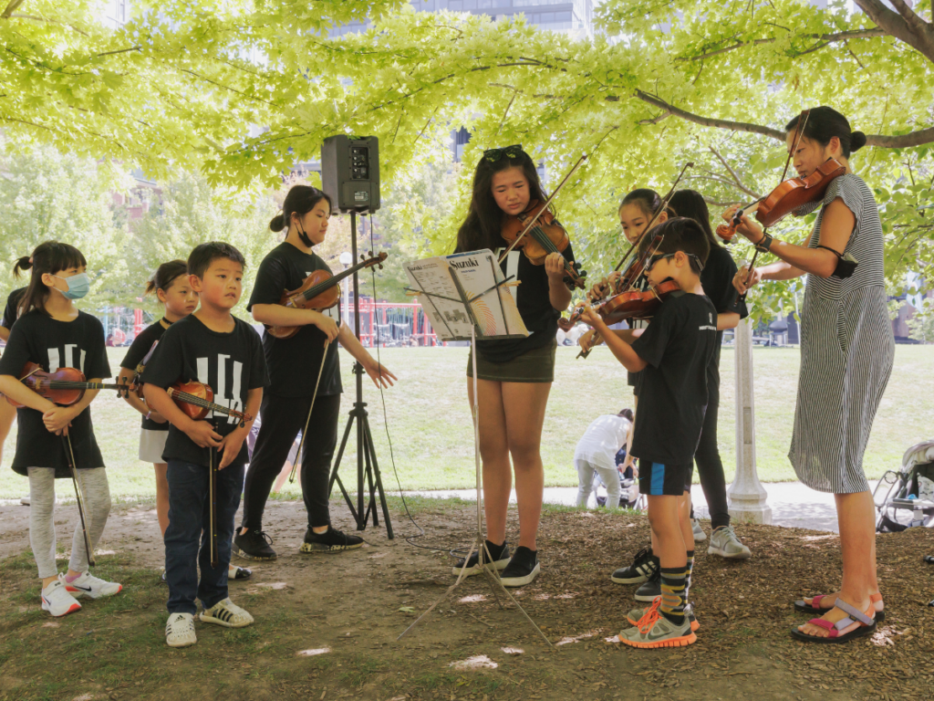 A group of students play violin outdoors, standing around a music stand.