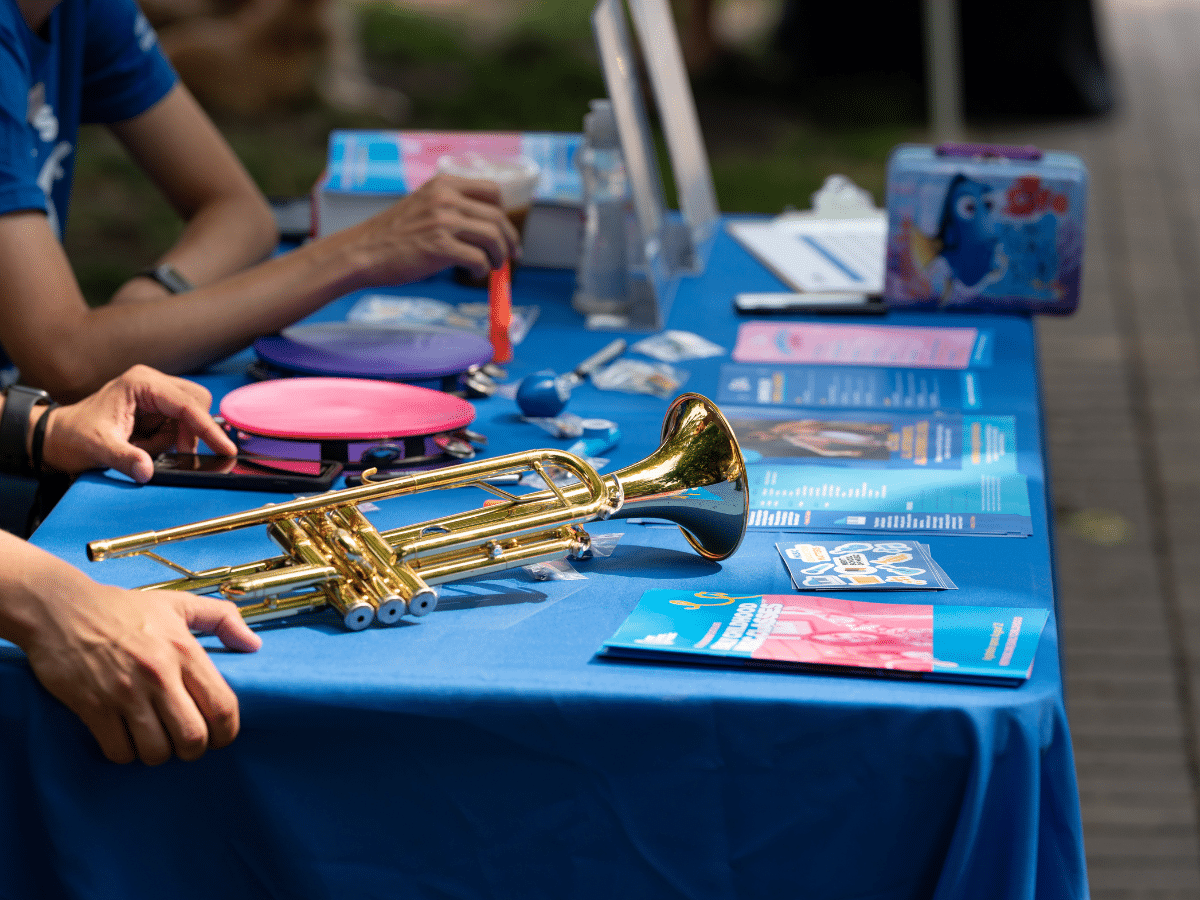 A close up of a table with a blue tablecloth at an open house. On the table is a trumpet, a tambourine, and flyers.