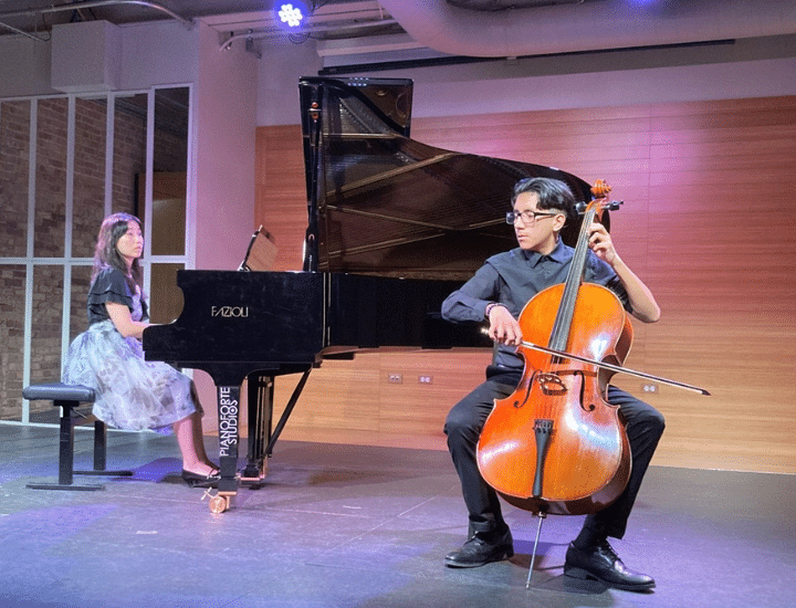 A student plays cello onstage with an accompanist at the piano.