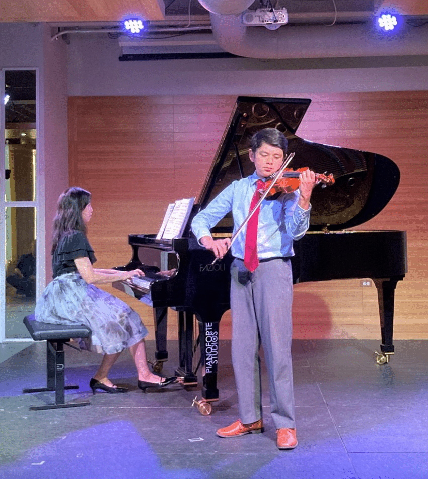 A student plays violin on stage, with an accompanist playing the piano.