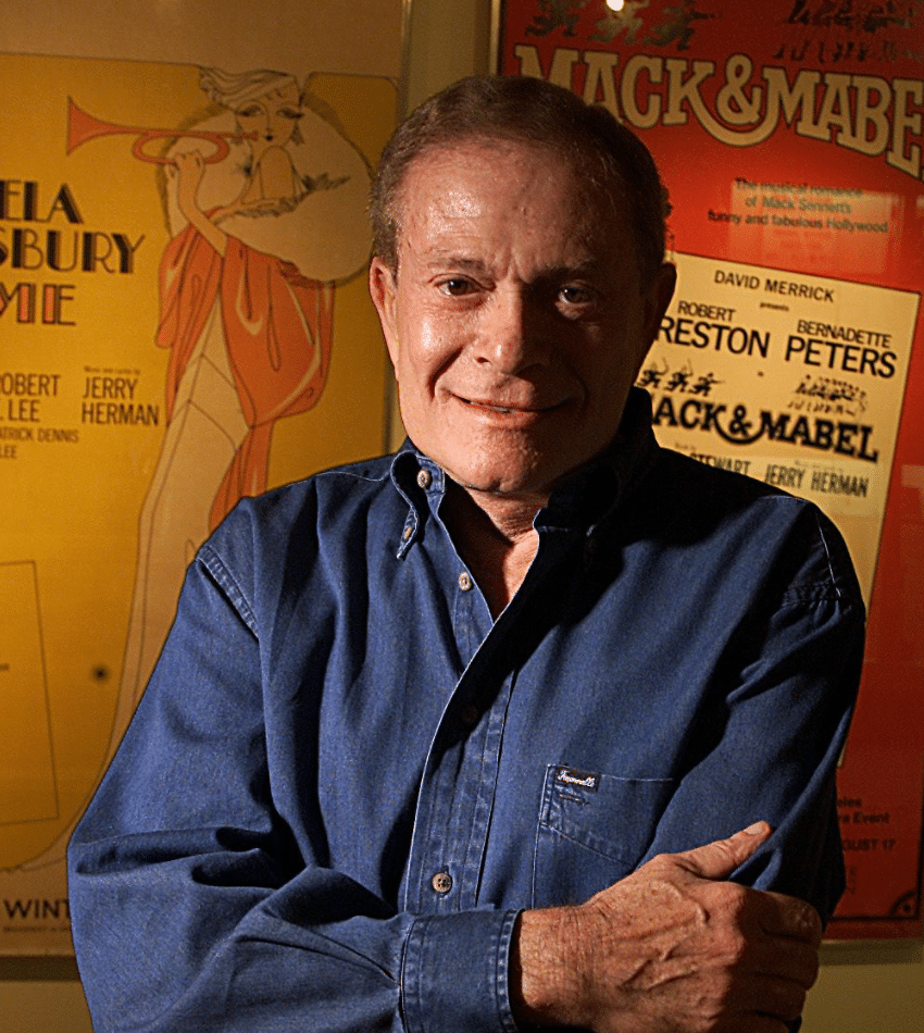 Jerry Herman poses with his arms crossed in front of posters of several of the musicals he composed.