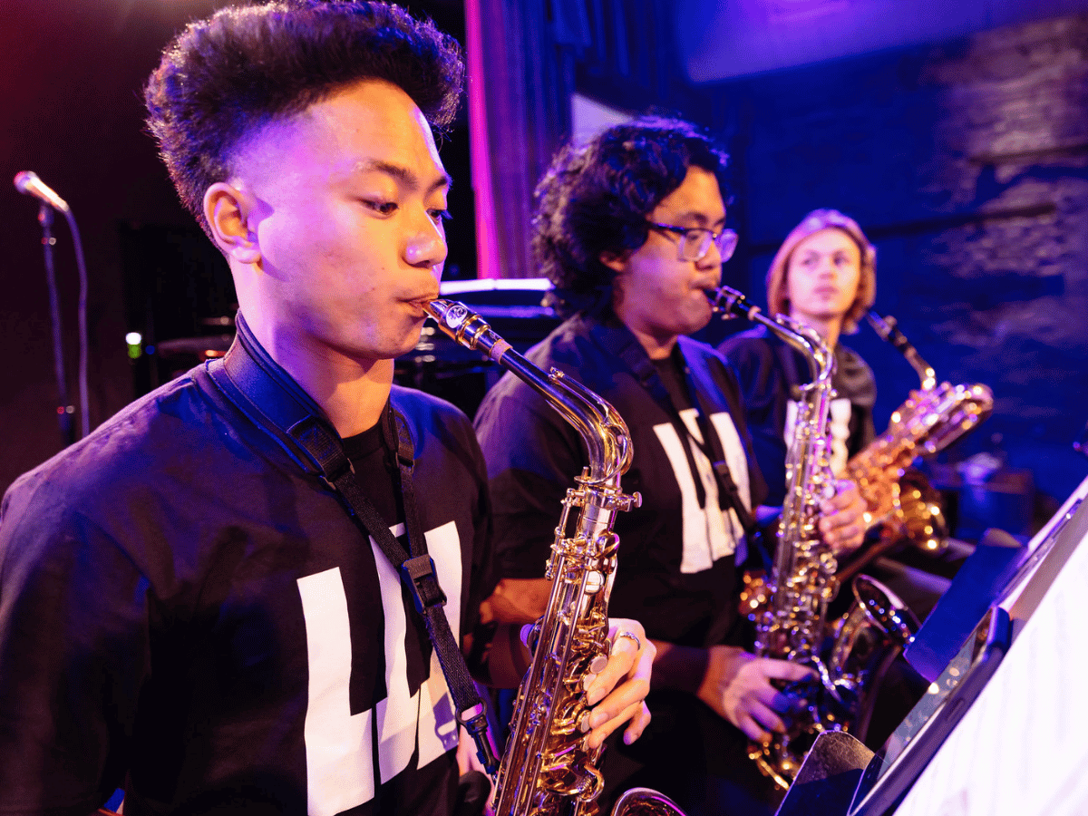 Three students play saxophone onstage under purple stage lights.