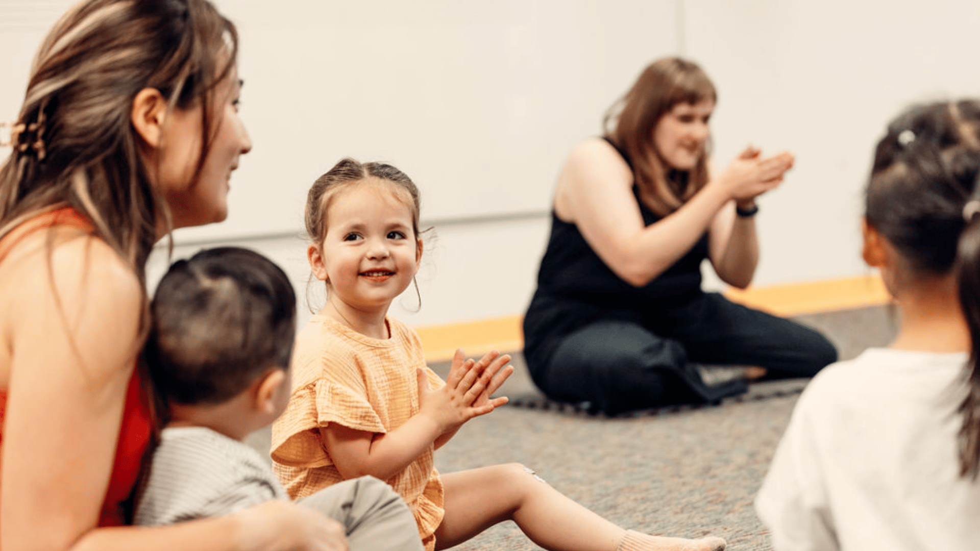 Several small children sit on the floor. A parent holds a baby in her lap, and the teacher is in the background.