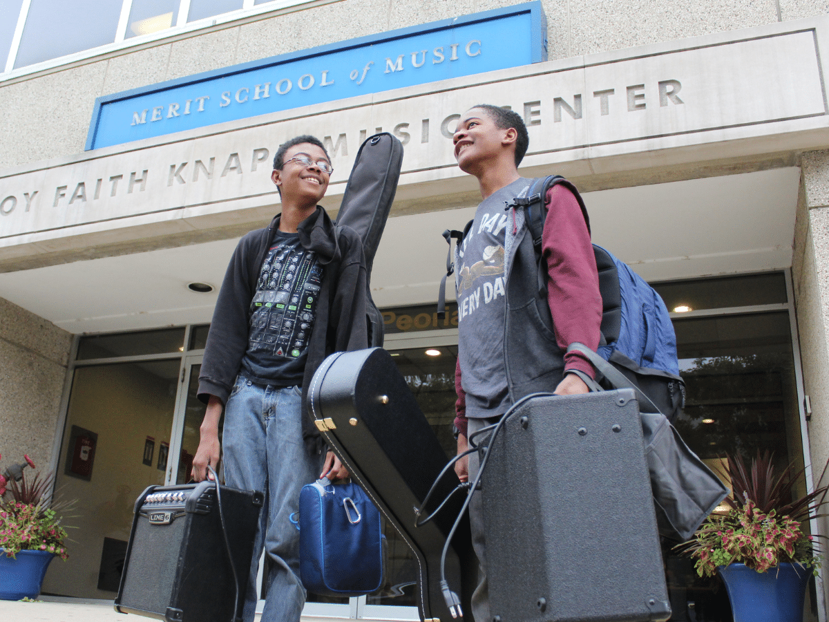Two students stand in front of Merit School of Music carrying guitars and amplifiers.