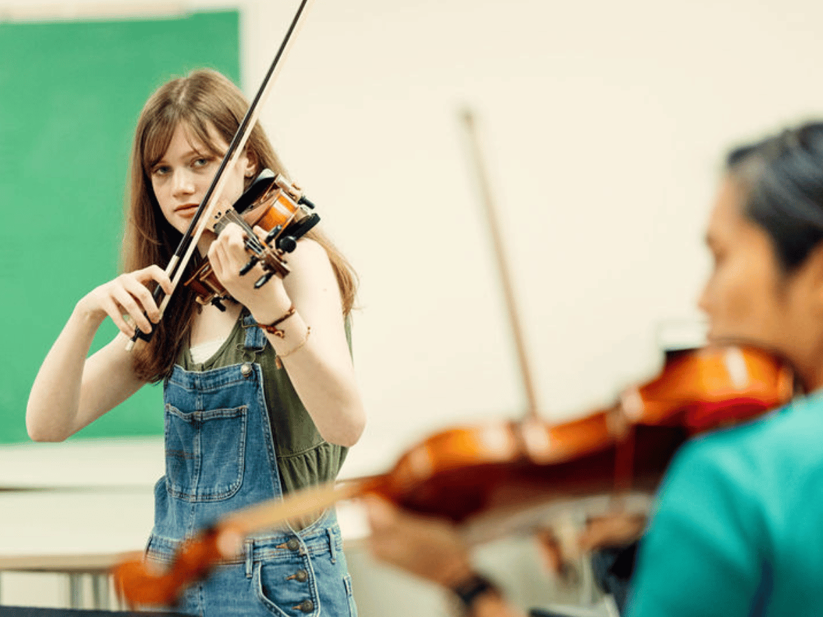 A student plays violin. Blurred in the foreground, her teacher stands facing her, holding a violin.
