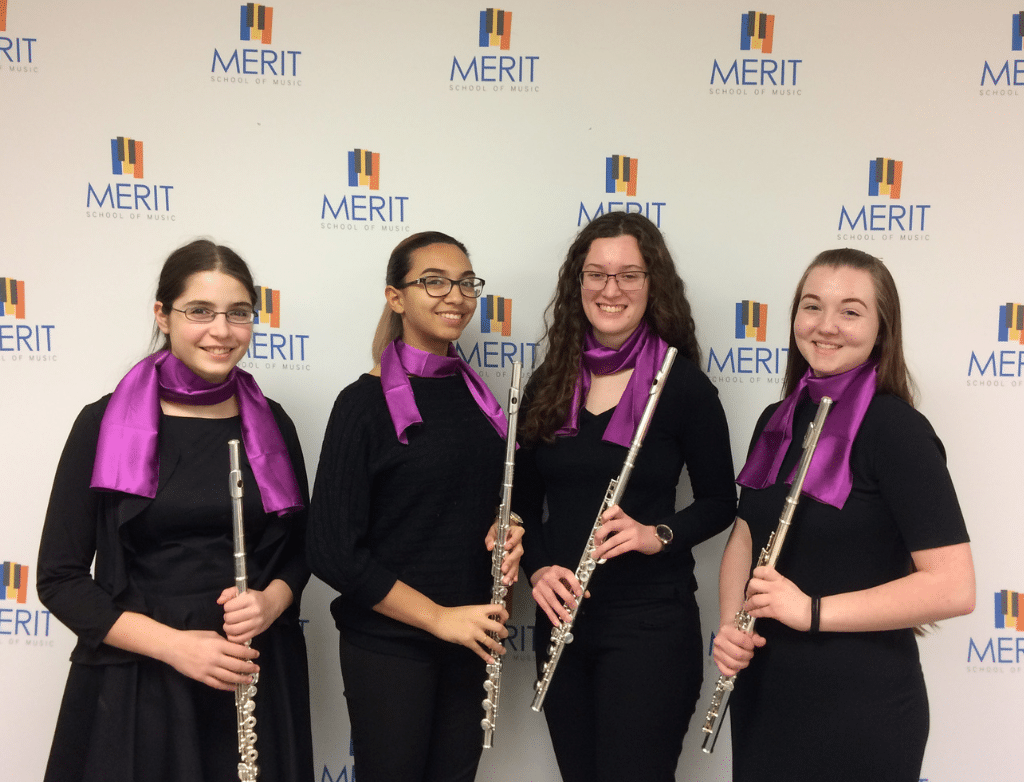 The 2016-2017 Purple Hippopotami pose together in front of a Merit School of Music step and repeat holding their flutes.