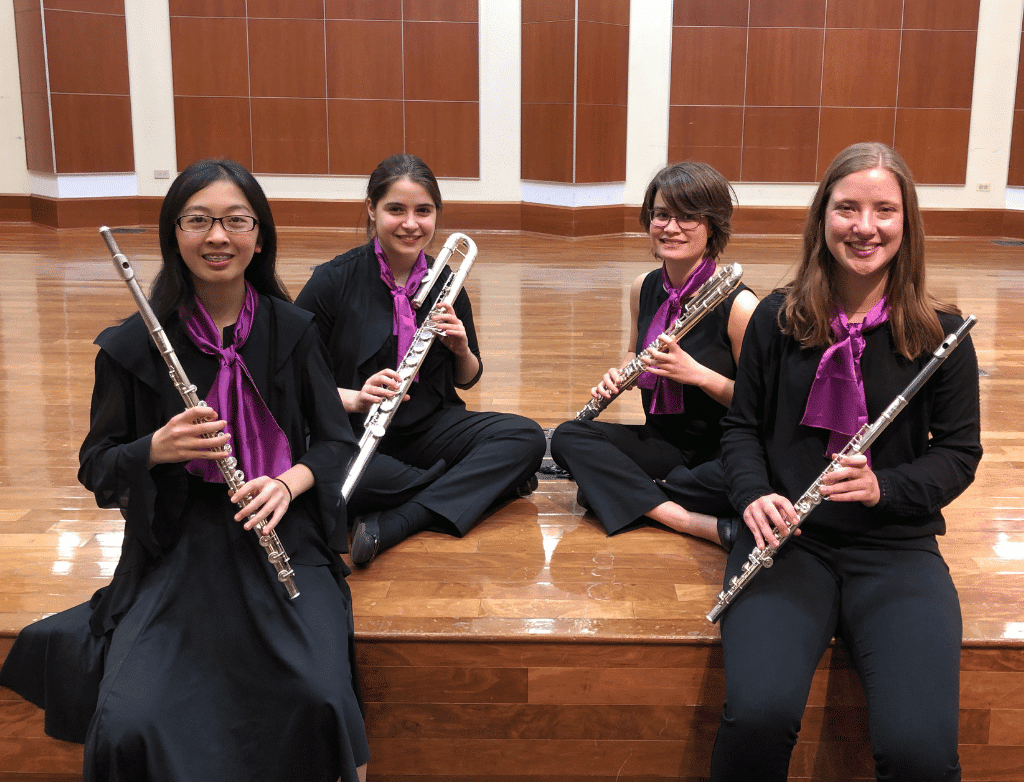 The 2019-2020 Purple Hippopotami sit casually on the stage of Gottlieb Hall, holding their flutes.
