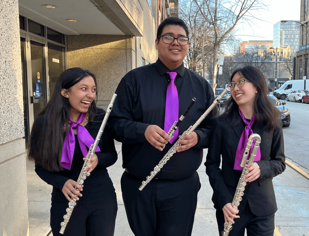 The 2022-2023 Purple Hippopotami laugh, holding their flutes and walking in front of the Joy Faith Knapp Music Center.