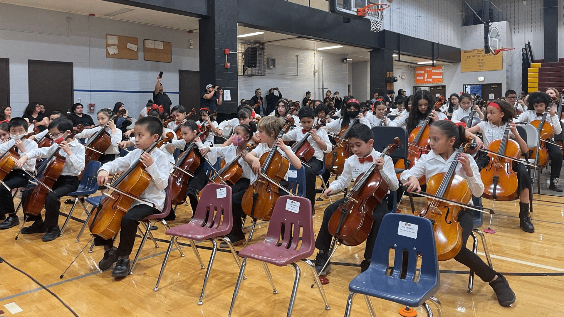 A large group of children playing cello in a gym.