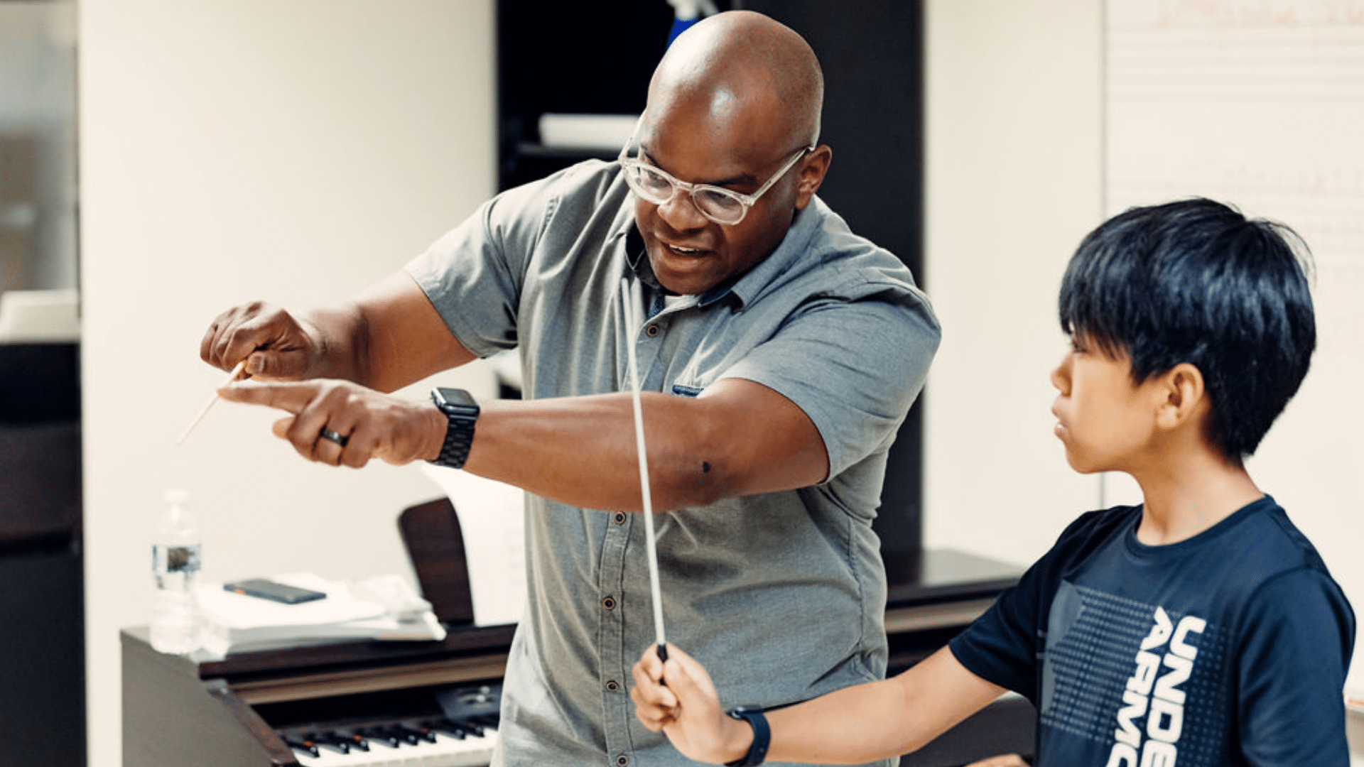 A student holds a baton, learning how to conduct from Steven Gooden, who is holding a baton and demonstrating.