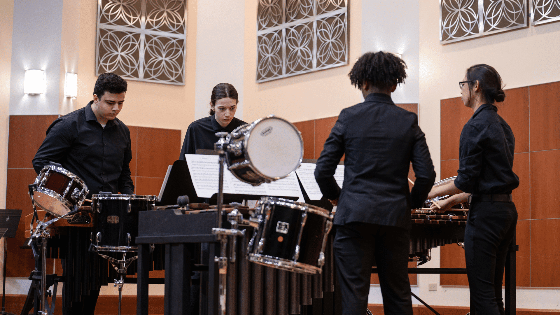 Four students in a percussion ensemble play various drums and percussion instruments.