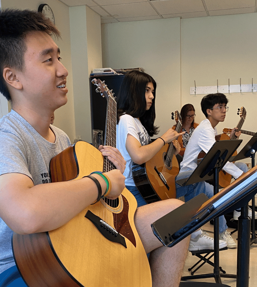 Three guitar students look toward the front of the classroom, smiling.