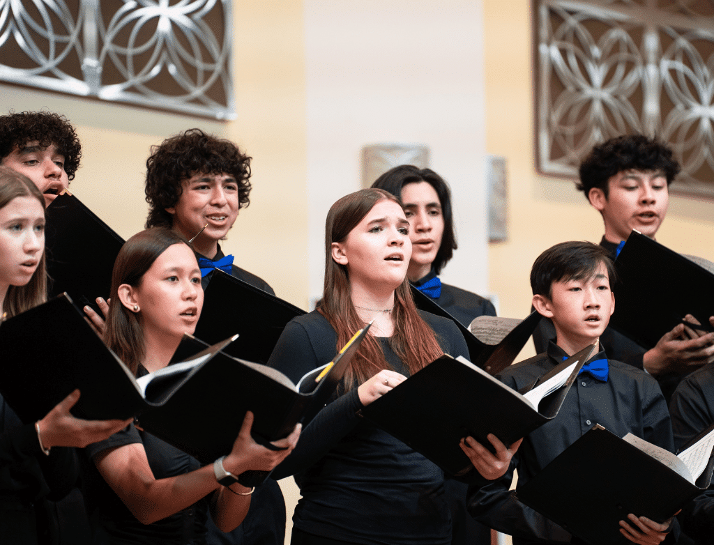 Students in a choir sing onstage, holding binders with their sheet music.