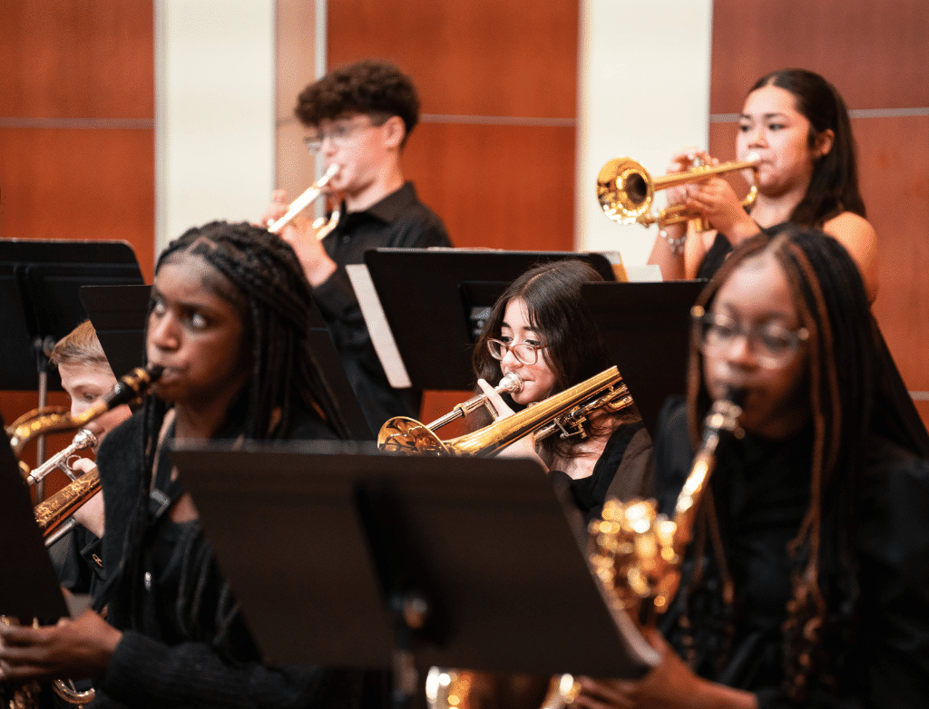 Students playing trumpets, trombones and saxophones perform onstage.