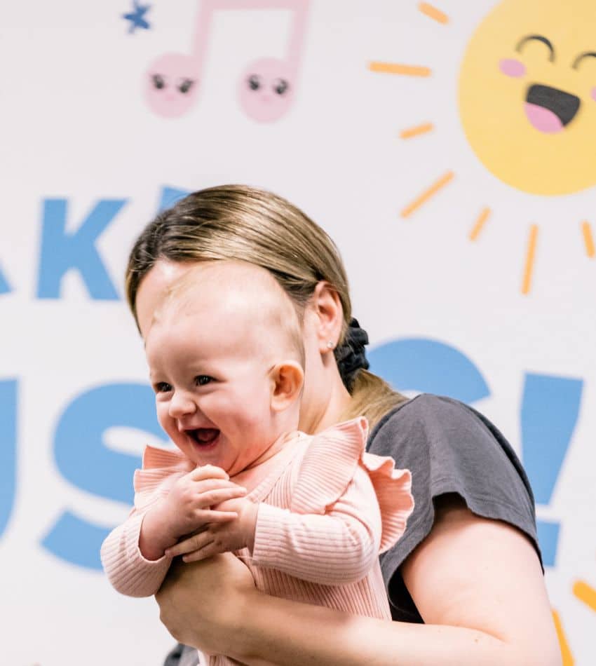 A parent holding a baby in front of a mural with a smiling sun and music notes