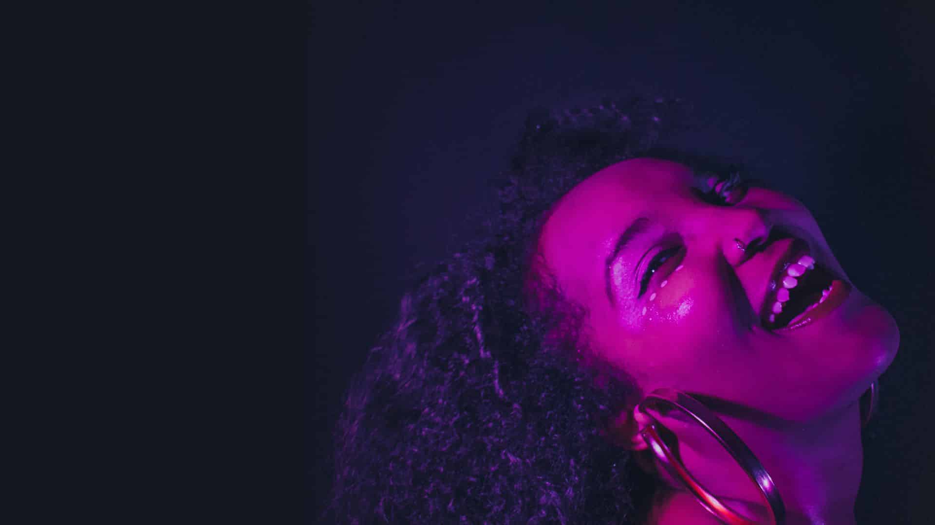 Alexis Lombre smiling with her head thrown back against a dark background. There is a bright purple light shining on her