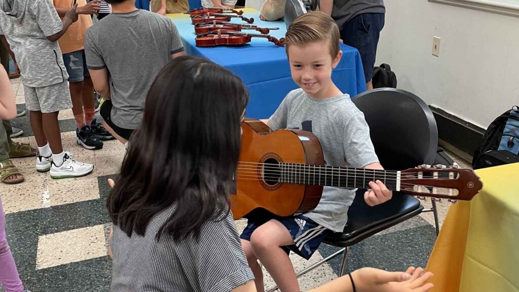 A child smiles holding a guitar while a teen helps him hold it correctly.