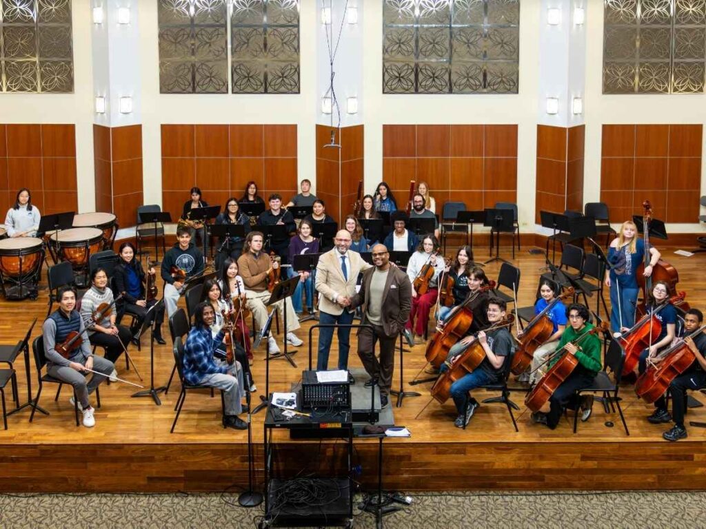 Merit Philharmonic sit onstage with their instruments, while Enrique Mazzola and Steven Gooden shake hands.