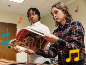 A student holding mallets stands next to his teacher, looking at a book together.