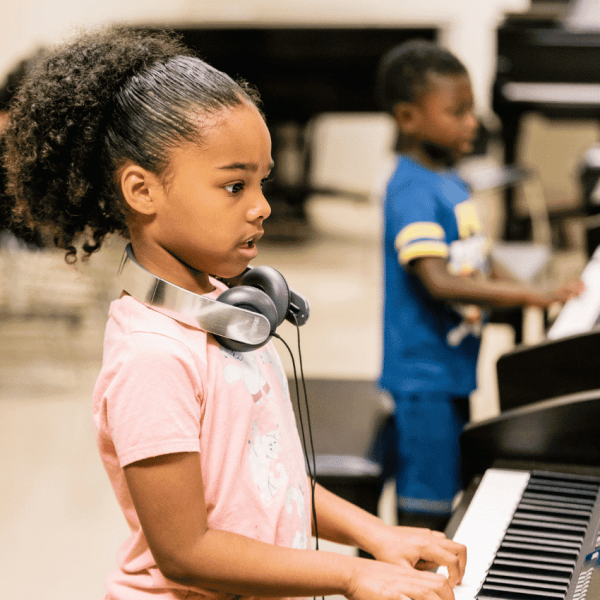 A young student plays keyboard standing up with a pair of headphones around her neck. Another student is blurred in the background.